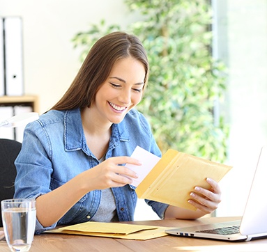 Woman in denim shirt smiling opening a yellow envelop at her work desk